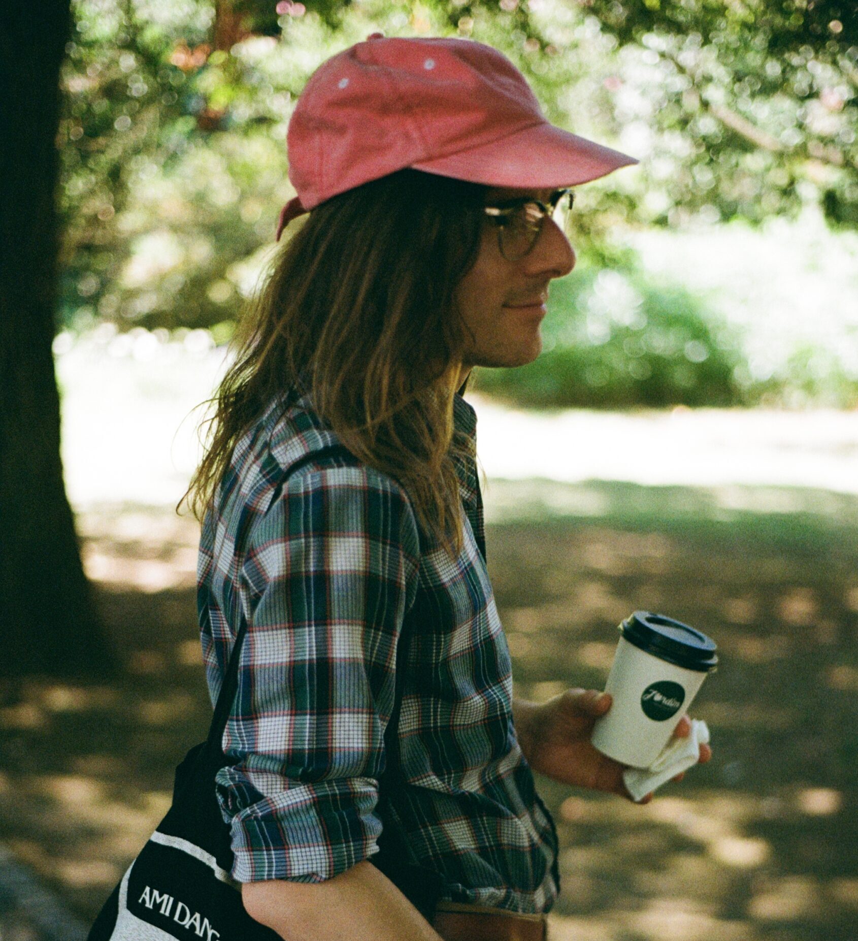 A man stands in the shade in a sunny park, he is wearing a baseball cap, holding a coffee, and a tote bag