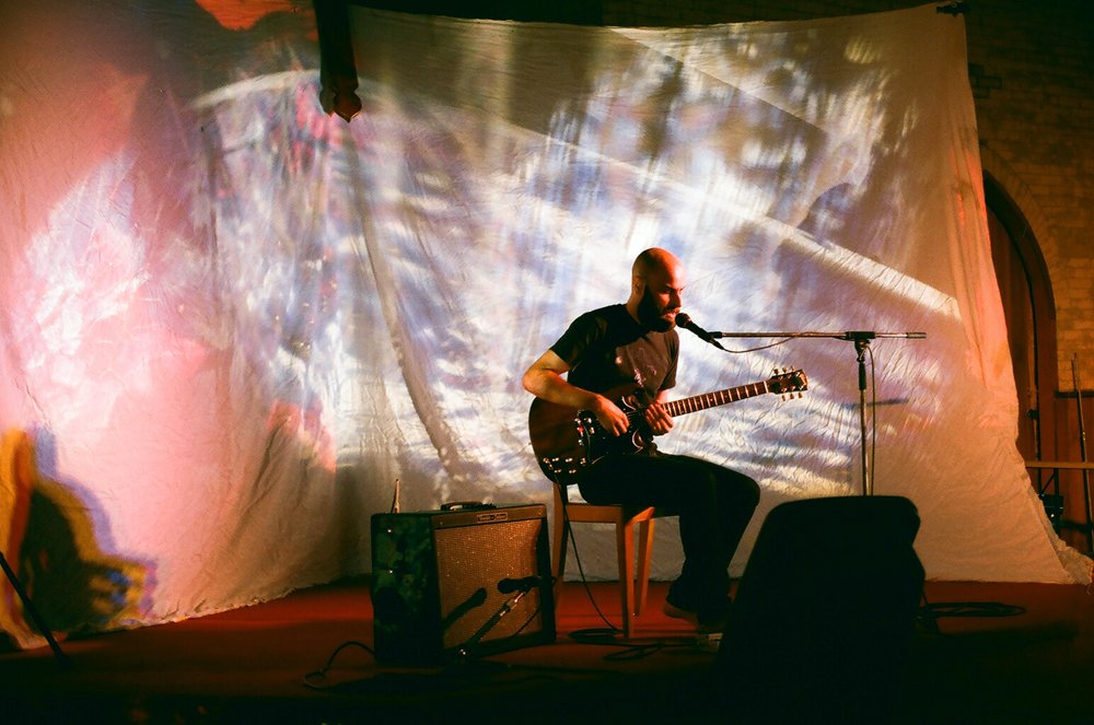 Man seated with guitar in front of a light show on a screen