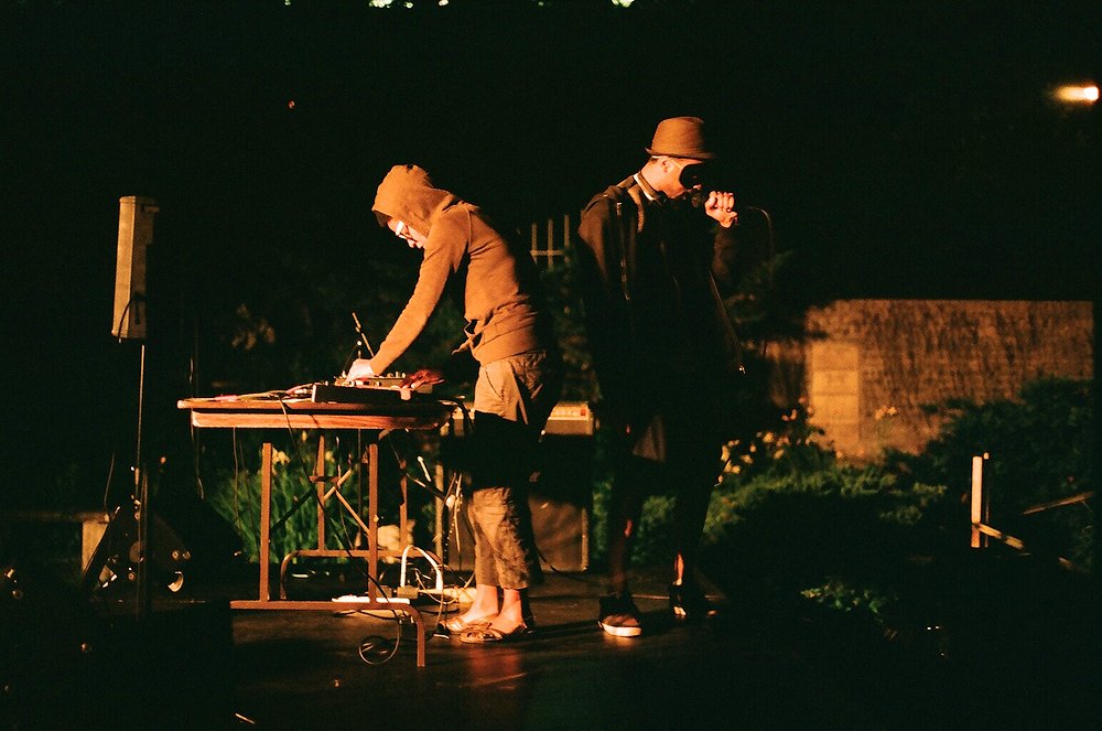A masked figure on the mic and an electronic musician. Nighttime, outdoors