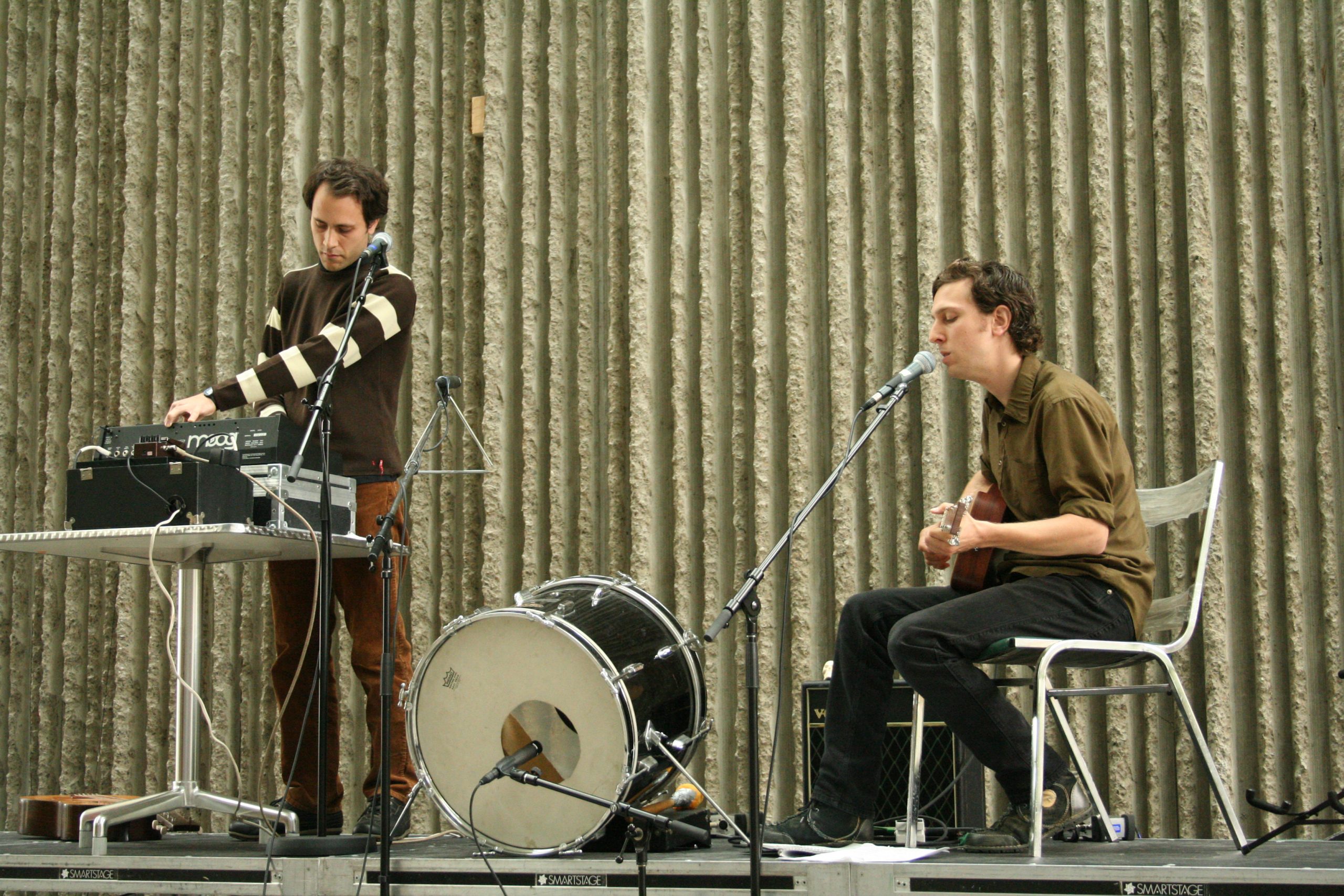 A singer/guitarist (seated) and electronic musician (standing) perform in front of a textured concrete wall