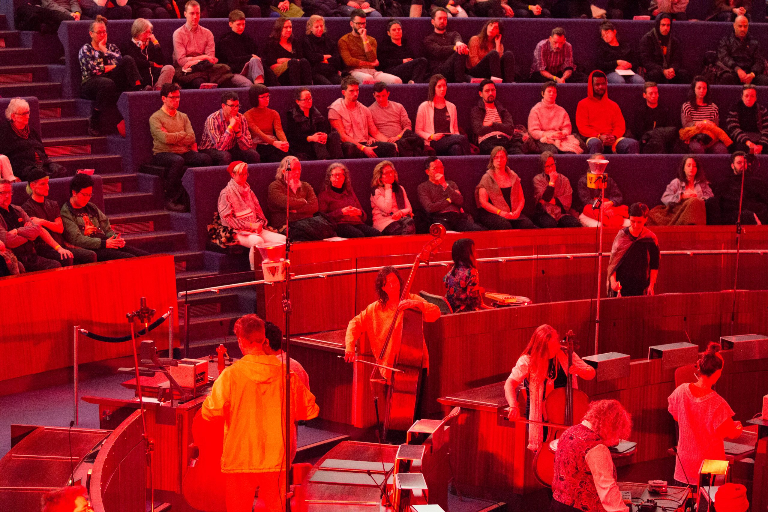 Musicians playing in an open indoor area bathed in red light surrounded by the audience