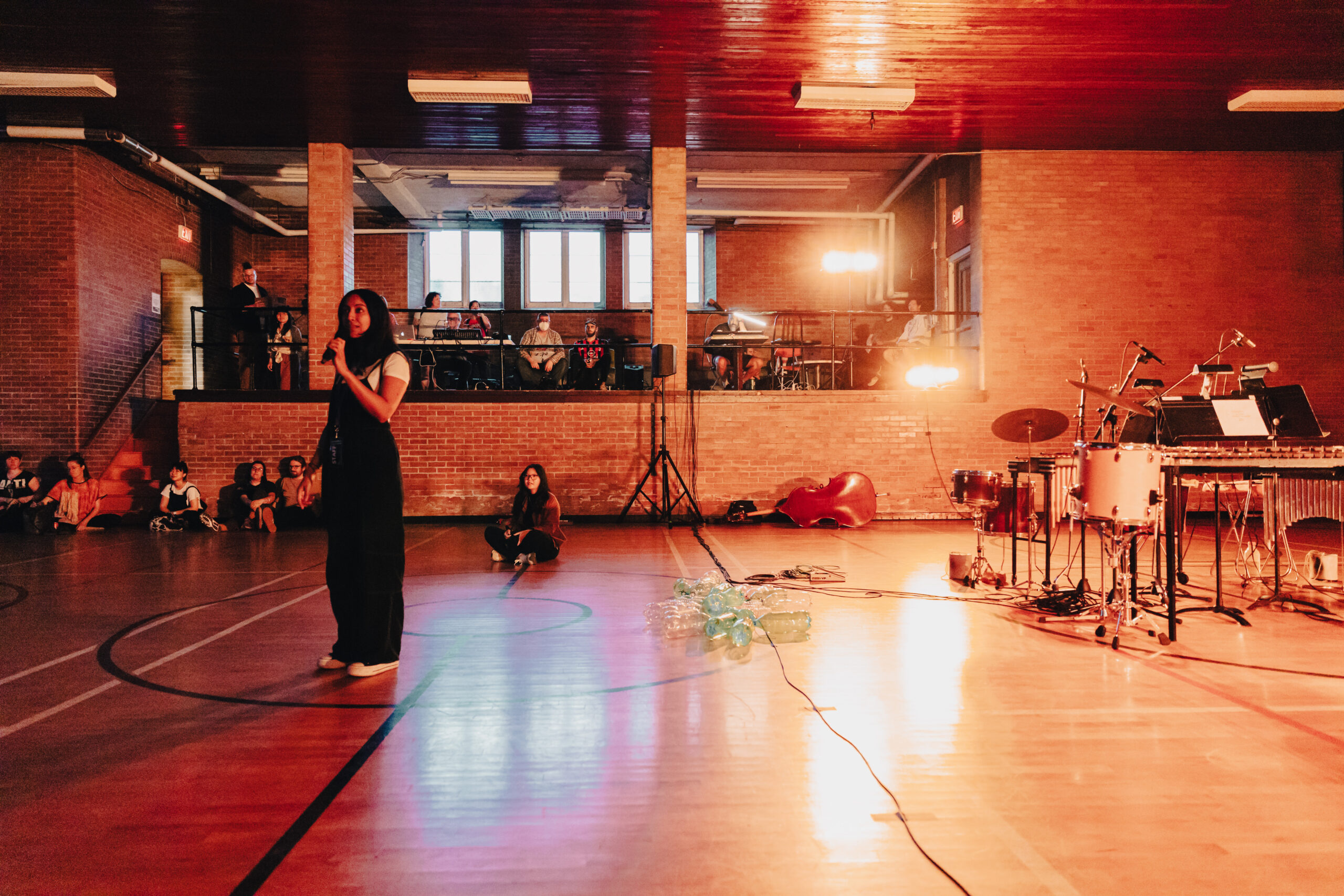 A wide shot of an indoor basketball court coloured by orange lights, a person stands in the foreground speaking on a microphone