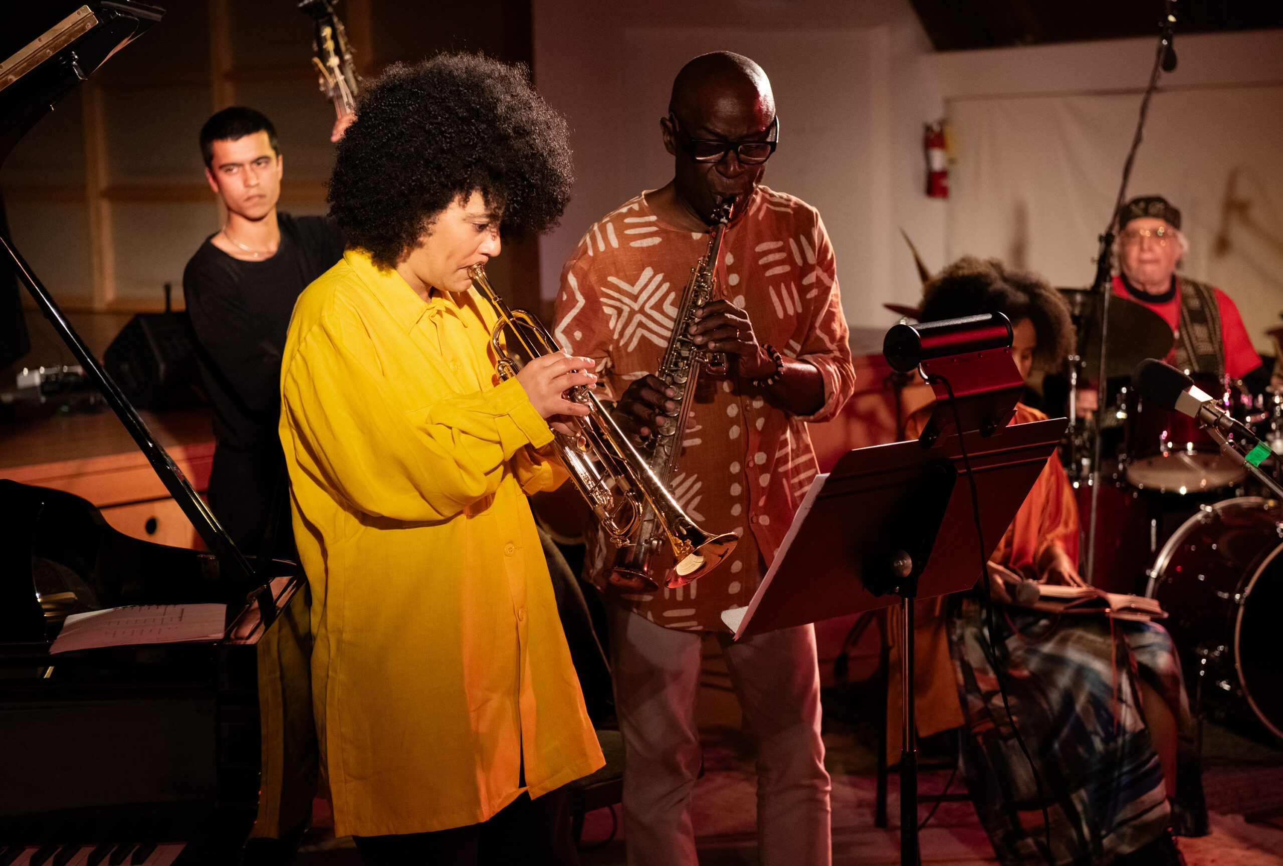 A group of musicians performing together, a trumpet player in a bright yellow shirt beside an alto sax player in a patterned shirt, both reading off a music in the foreground.