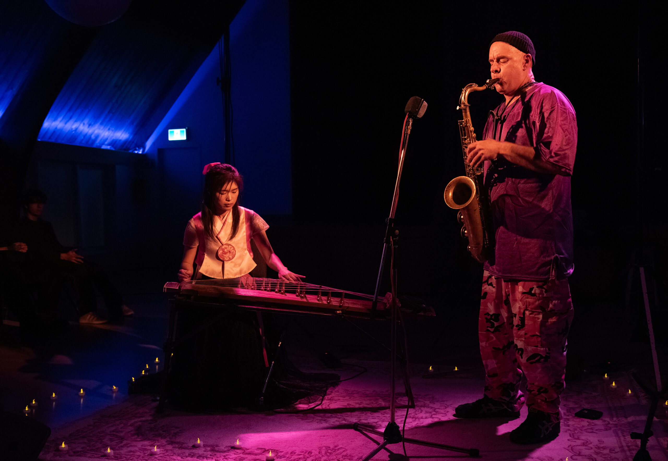 Two musicians perform together in dramatic magenta lighting, on standing, one seated